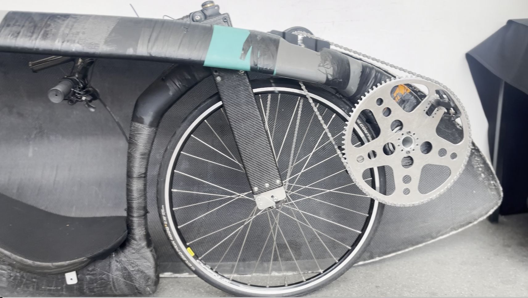 Close-up of an aerodynamic bicycle prototype with a carbon fibre body shell, exposed front wheel and large metal chainring, showing mechanical components and lightweight composite construction.