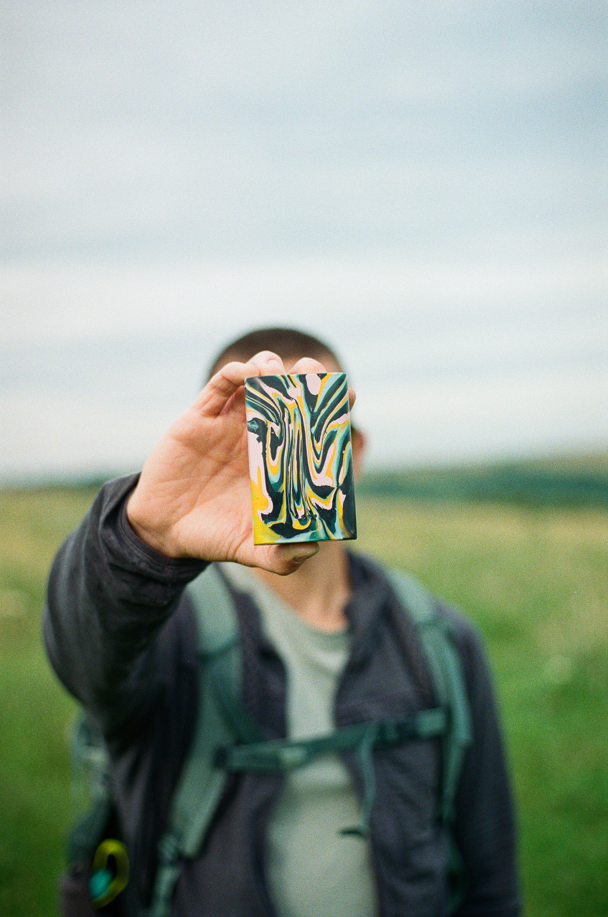 Person holding a small rectangular recycled plastic device with a marbled pattern of black, yellow and cream, photographed outdoors with the background softly blurred.