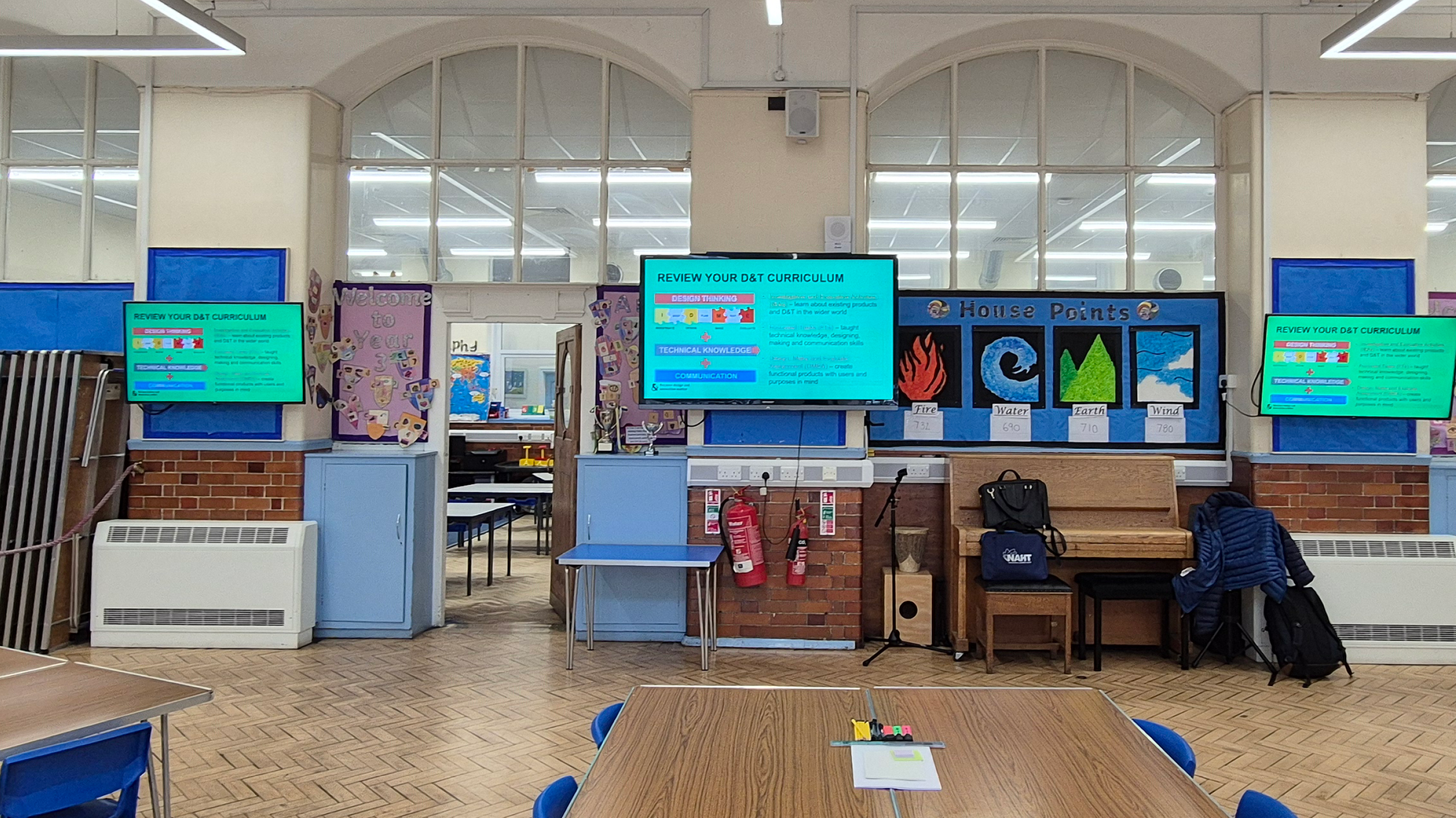 Classroom with screens on the wall, a piano against the back wall, tables and chairs