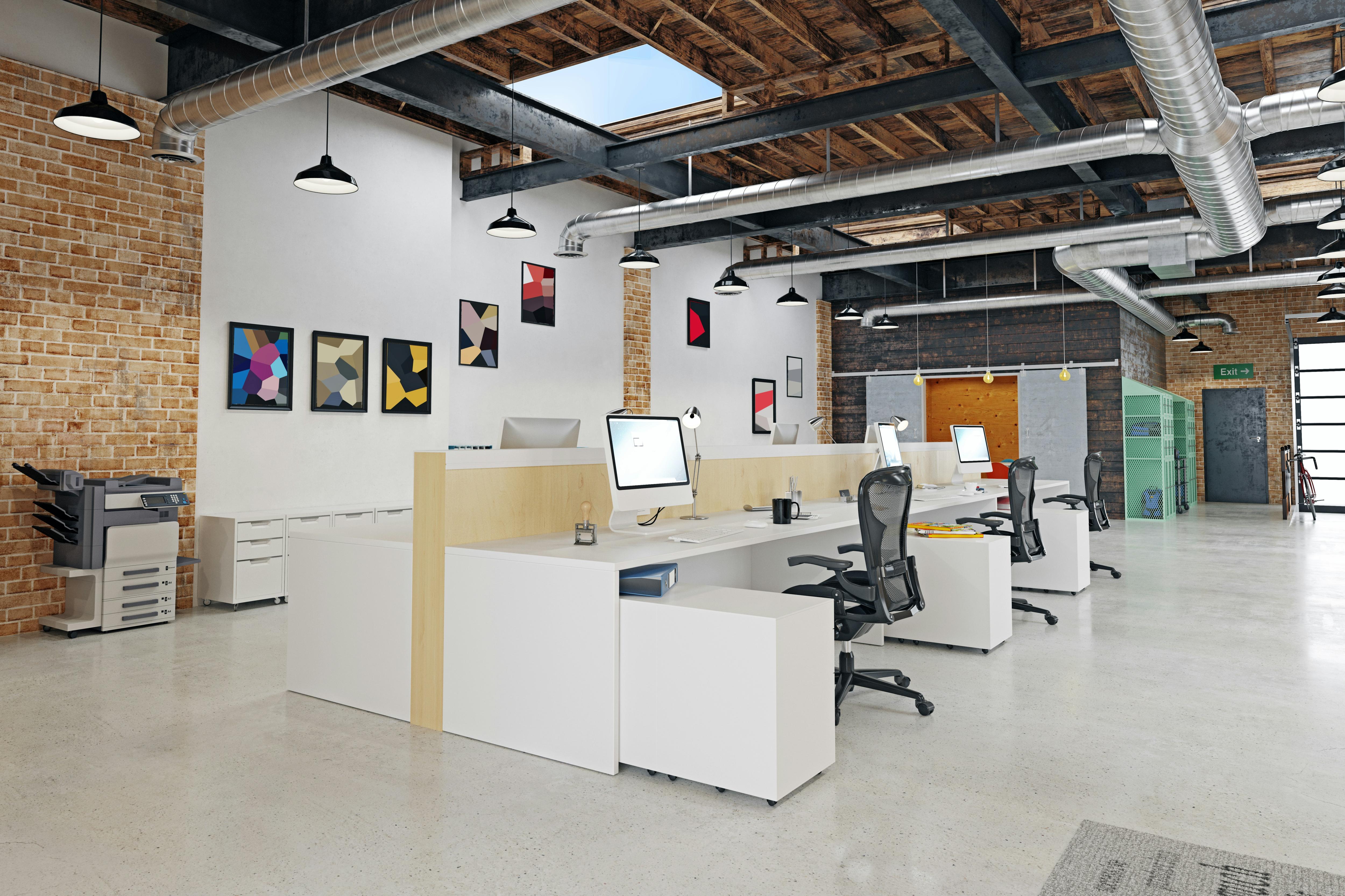 Modern open-plan office with desks, computers, office chairs and exposed industrial ceiling pipes.