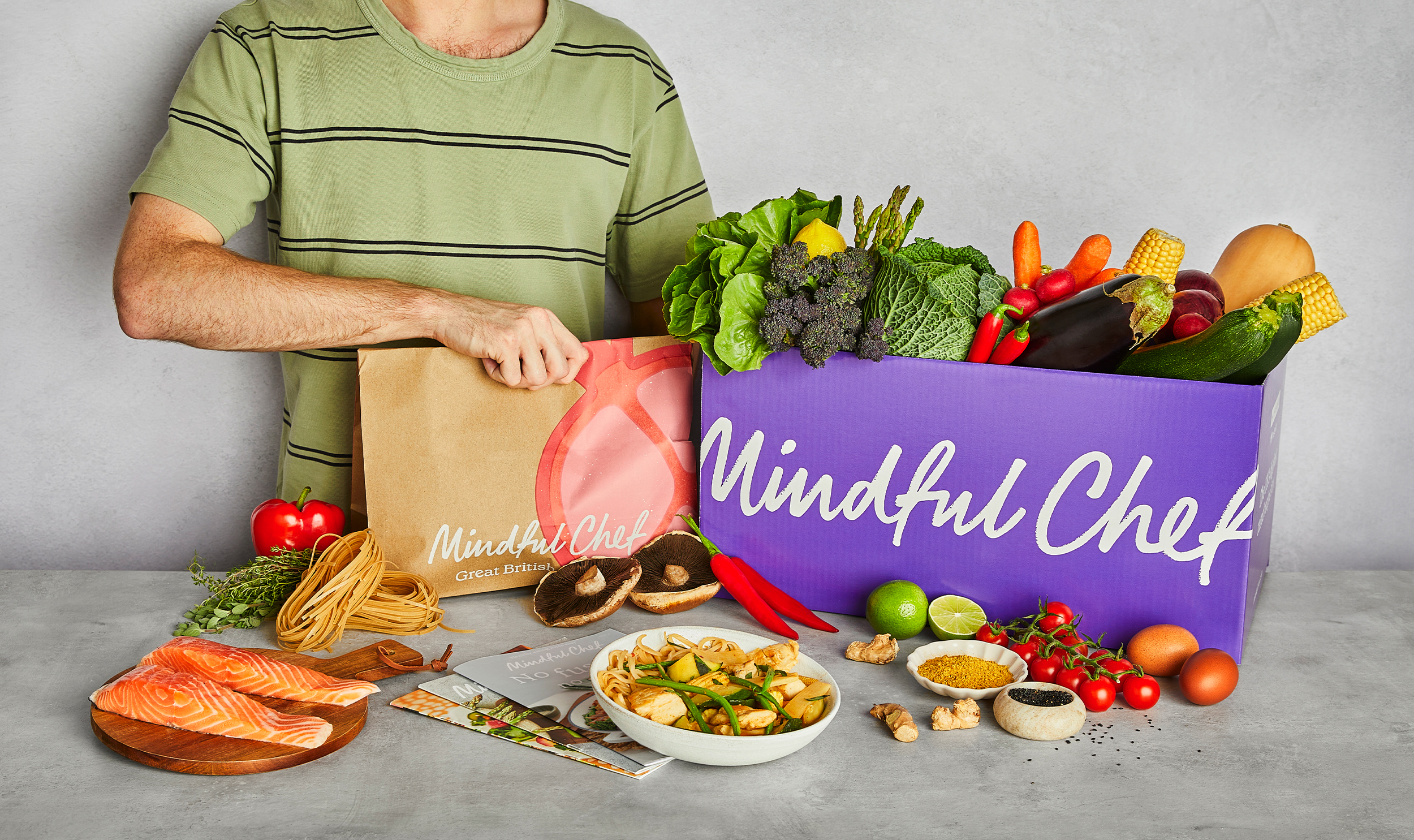 Person unpacking a Mindful Chef recipe box filled with fresh vegetables, salmon, pasta and ingredients for healthy home cooking.