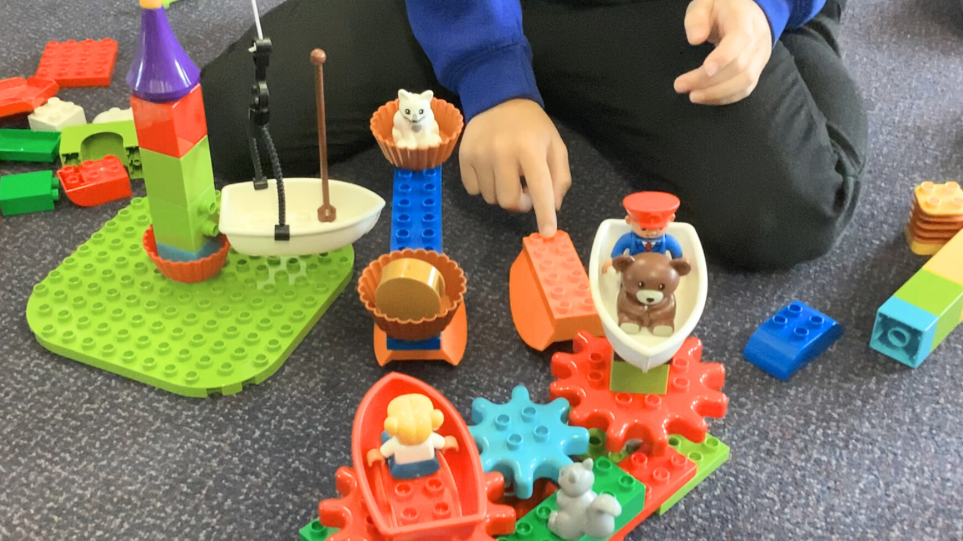 A young child builds and explores simple structures and mechanisms using colourful construction blocks and small figures on the floor.