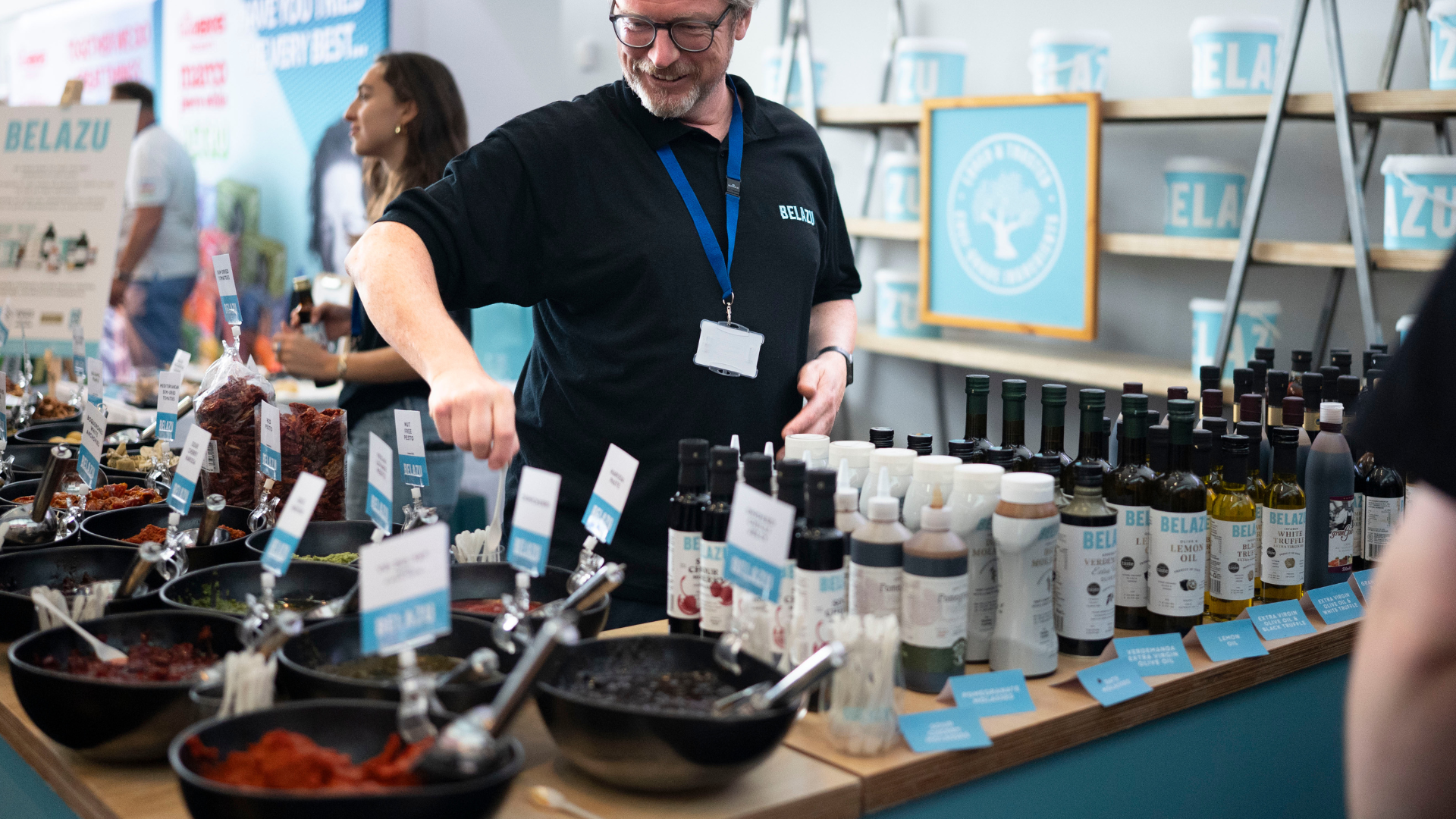 A Belazu team member serving samples at a tasting stand, with bowls of Mediterranean ingredients and bottles of oils and vinegars on display.