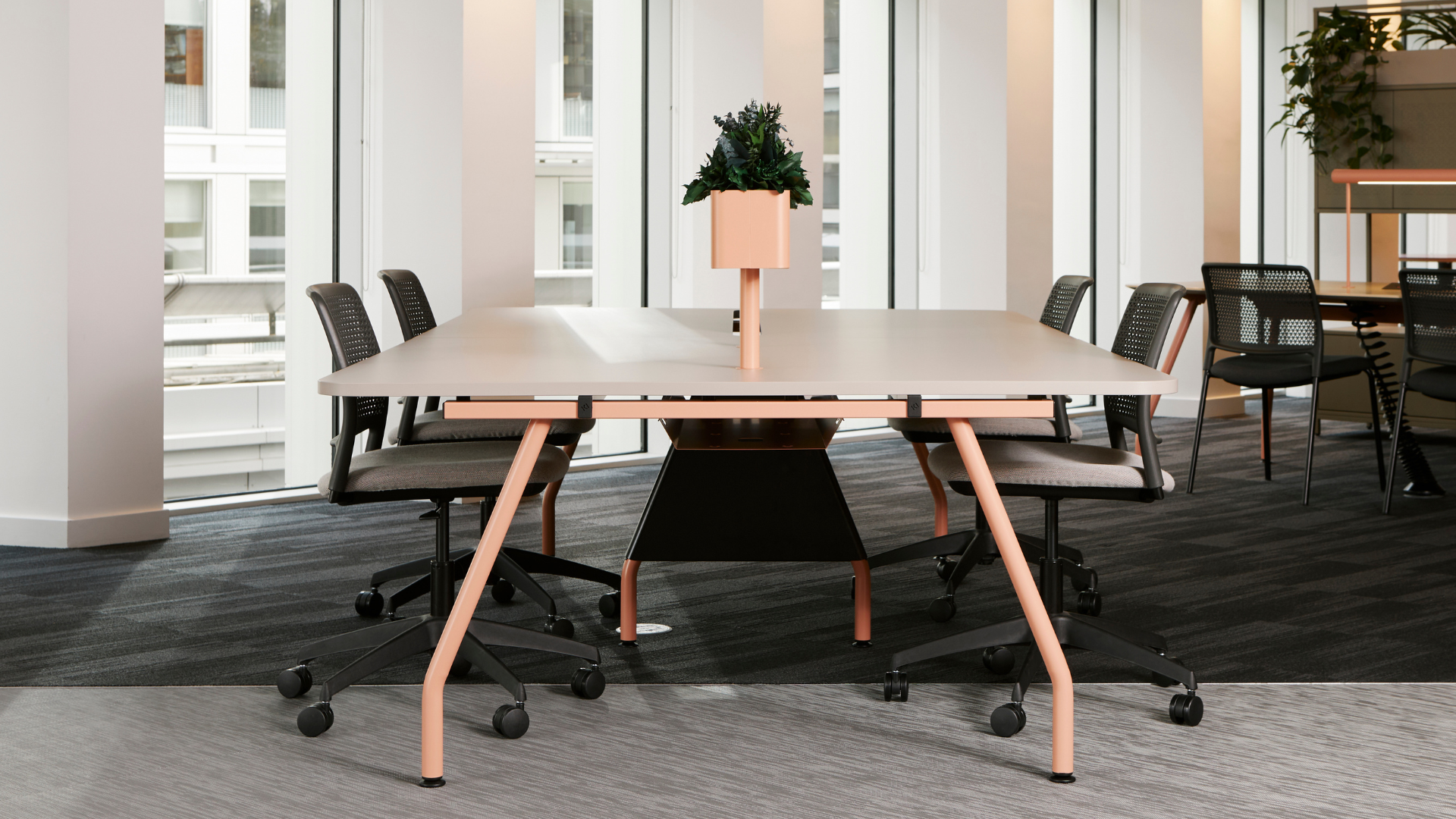 A modern office workspace with a large table, wheeled task chairs, and integrated lighting, demonstrating contract furniture design and ergonomics.