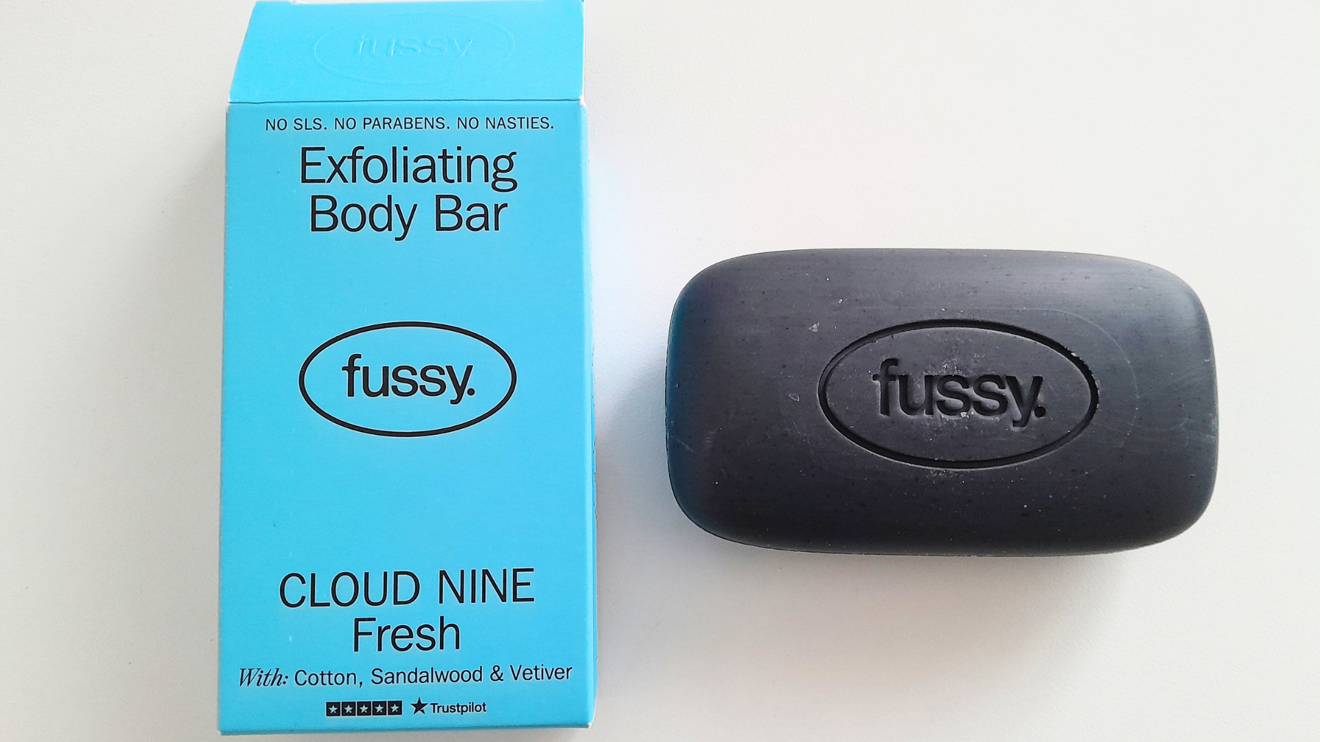 Fussy exfoliating body bar shown next to its blue cardboard packaging labelled “Cloud Nine Fresh,” with the black soap bar featuring an embossed fussy logo.