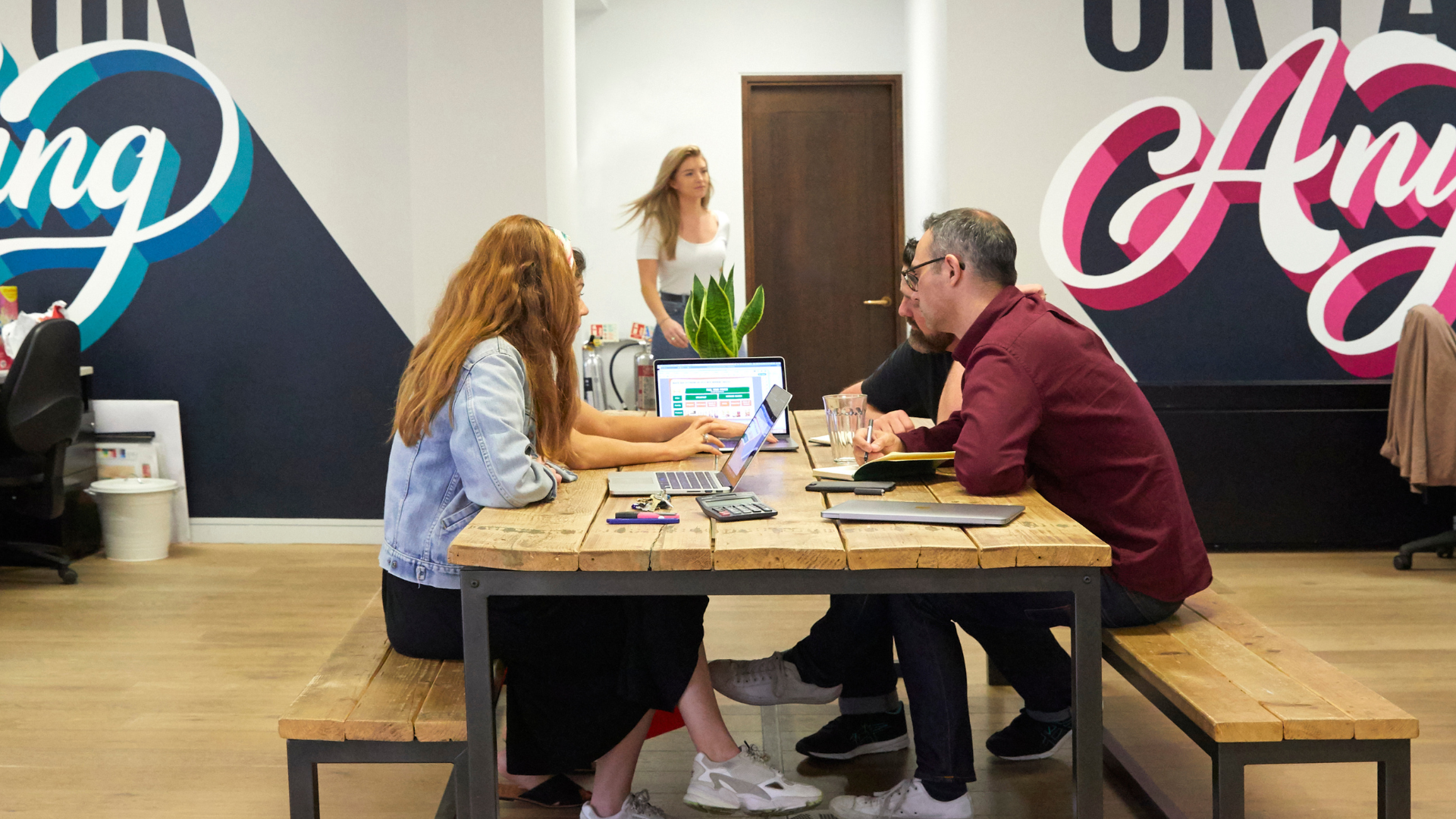Team working together around a table in a creative office space, using laptops and notebooks during a collaborative design and planning meeting.