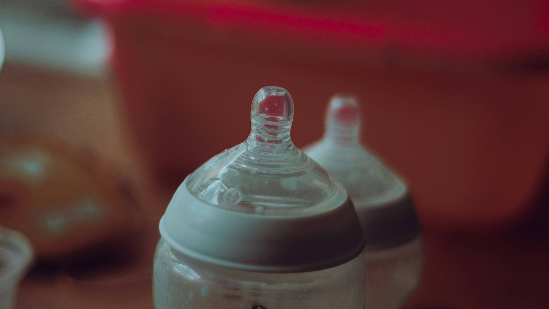 Two baby bottles with silicone nipples in soft focus, set against a warm red background.