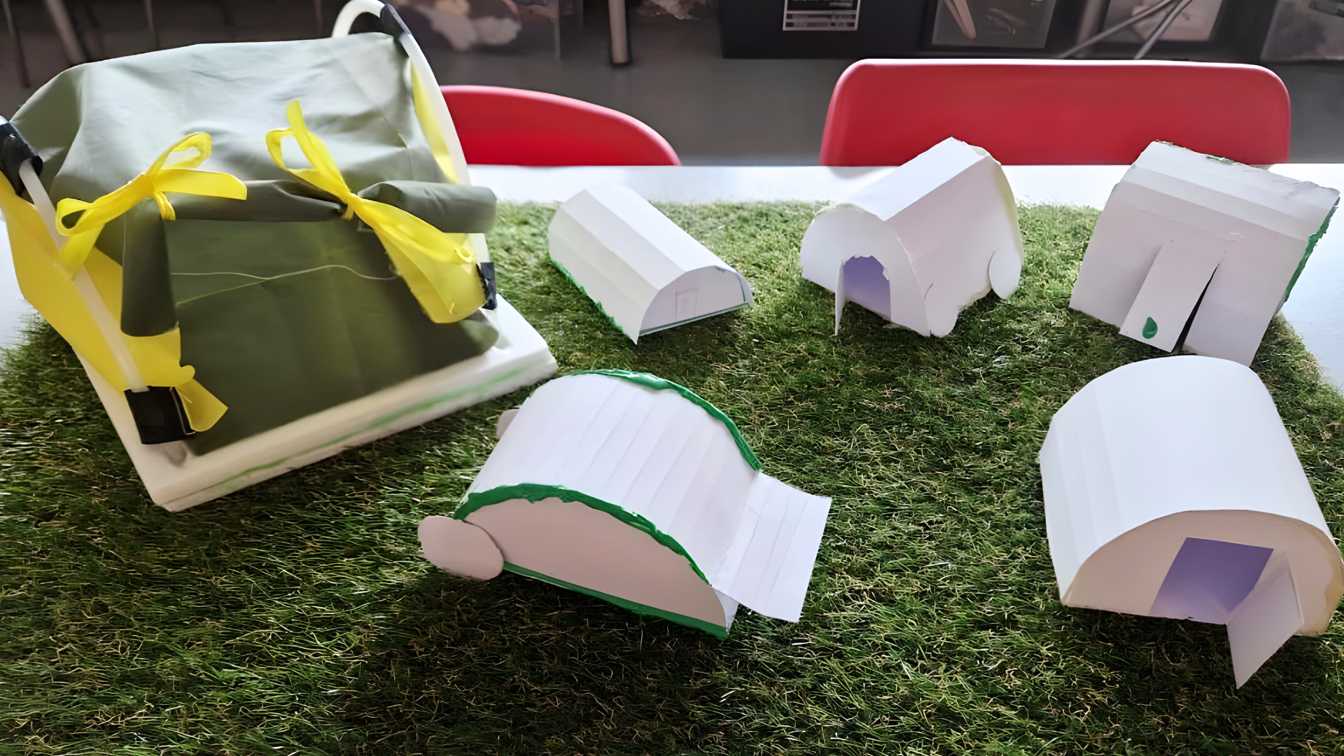 Early-stage short-term shelter models made from card and mixed materials, displayed on a grass-effect base.