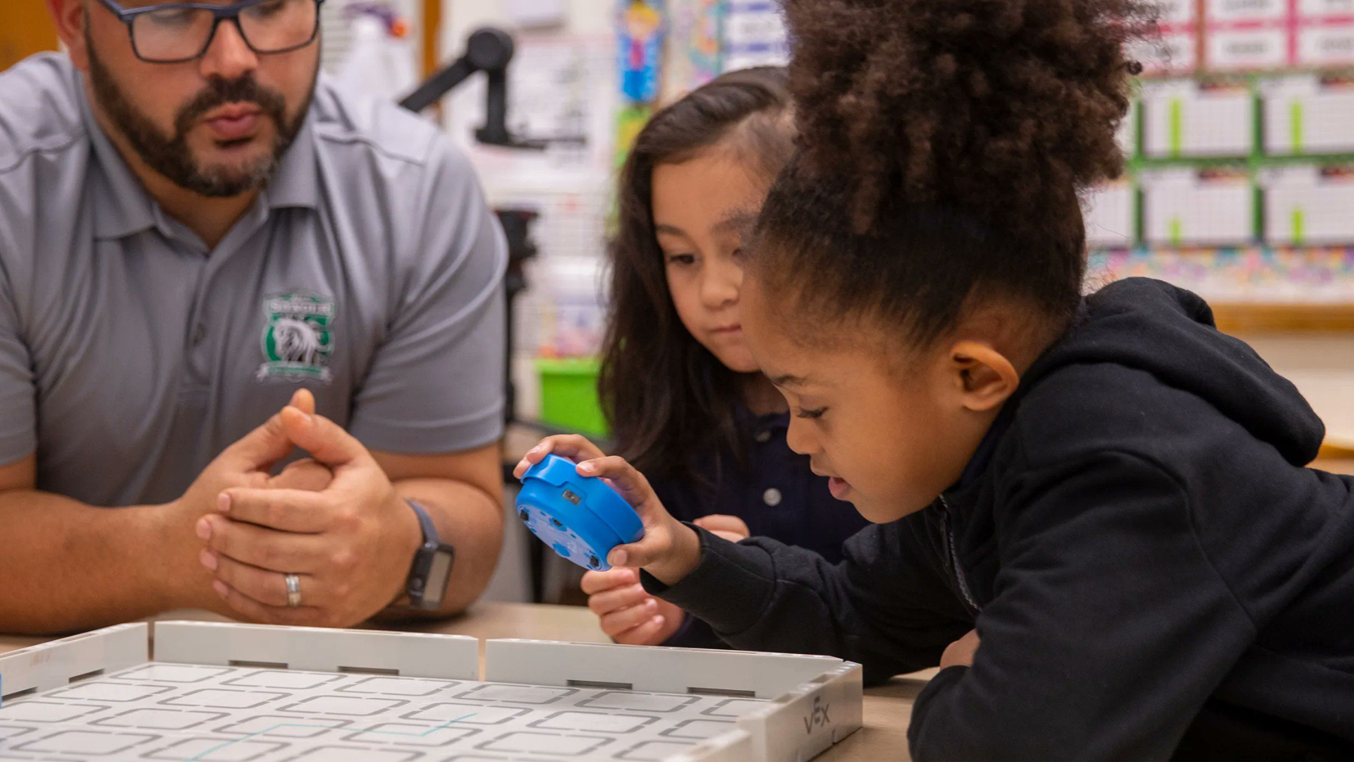 A teacher supports two primary pupils as they use a small programmable robot on a tabletop maze during a design and technology lesson.