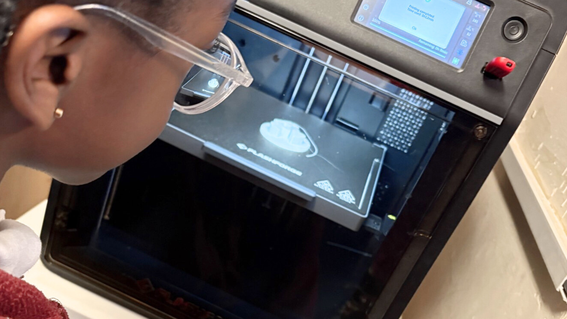 A student wearing safety glasses observes a 3D printer producing a component inside the machine.