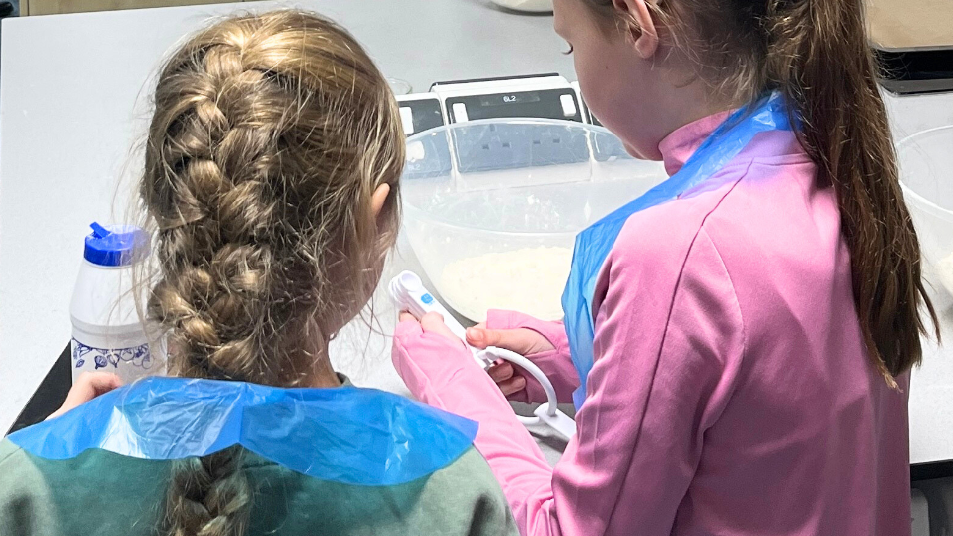 Two primary pupils wear aprons while measuring and mixing ingredients at a classroom workbench.