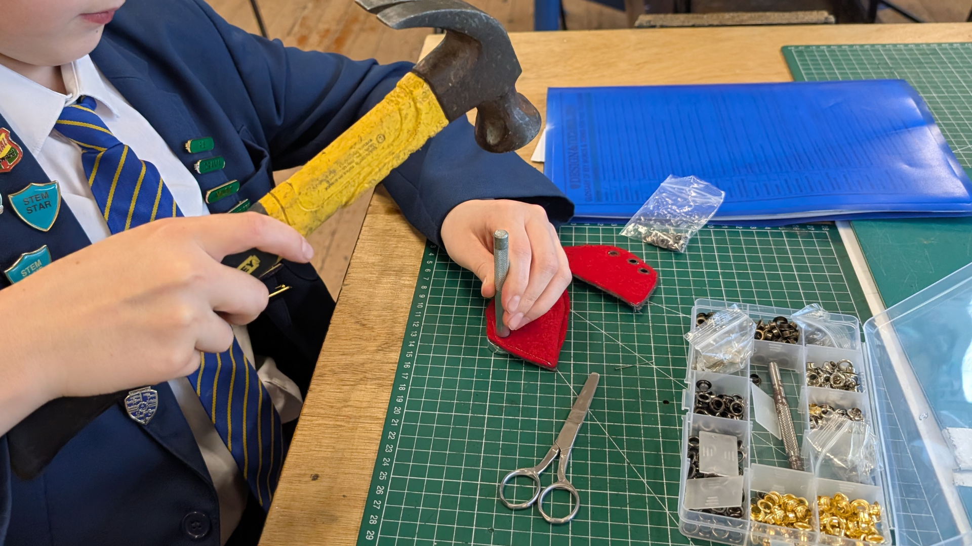 Student using a hammer and punch to fix metal fasteners into a red material piece on a cutting mat in a design and technology lesson.