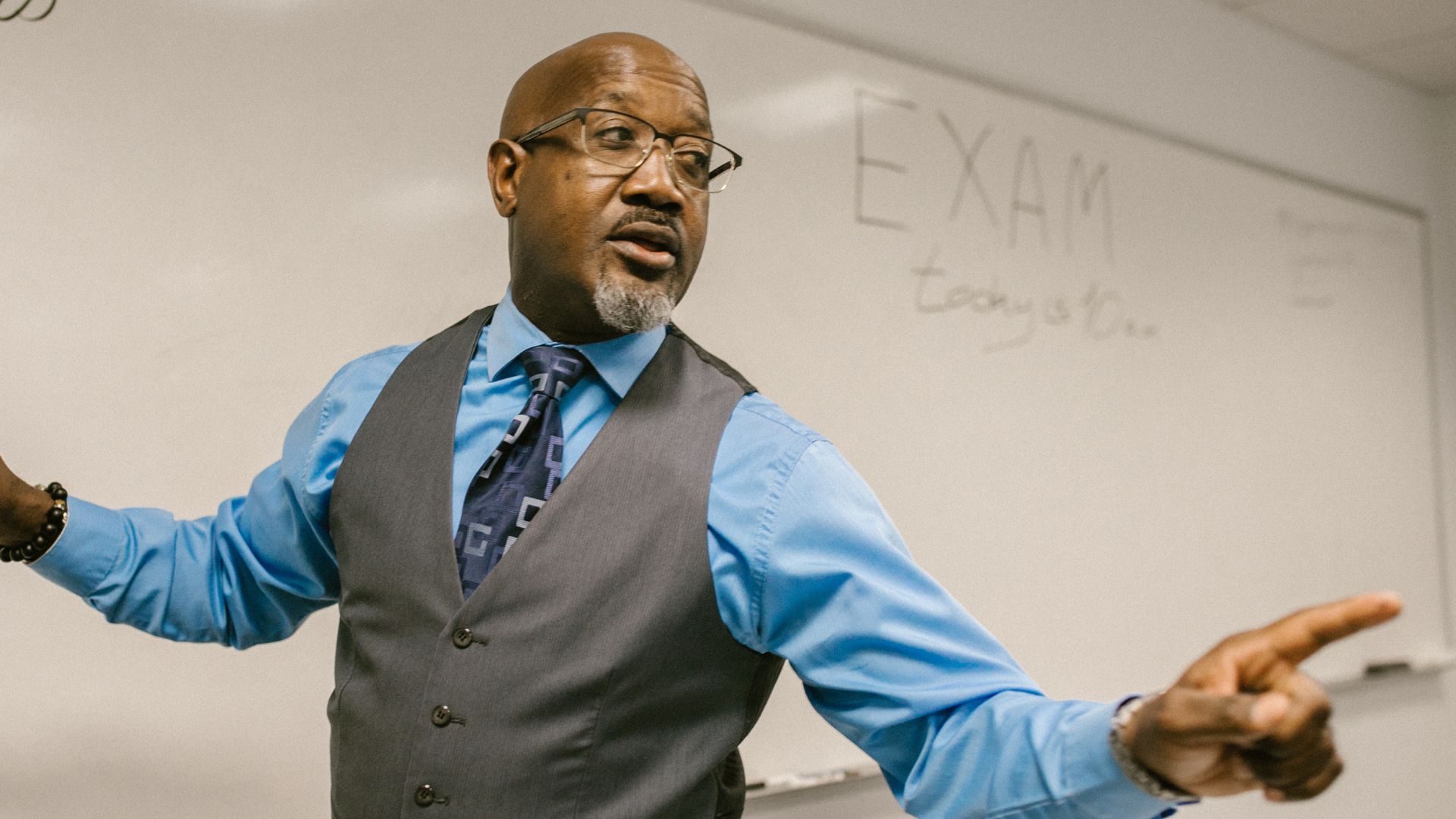An experienced teacher presenting to a group, gesturing while explaining a concept in front of a classroom whiteboard.