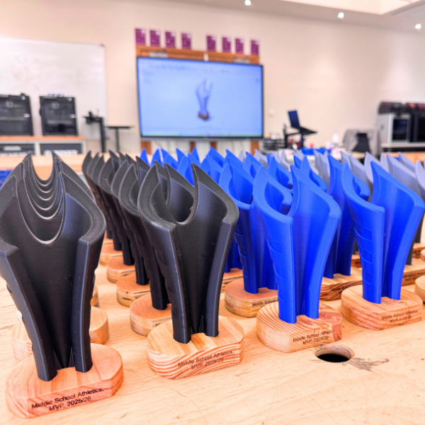Rows of student-designed 3D printed trophies in black and blue displayed on wooden bases in a classroom workshop setting