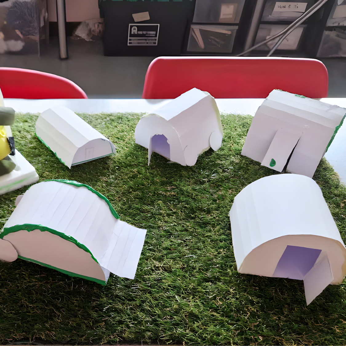 Range of student-made shelter prototype models constructed from card, exploring different forms and structures on a grass base in a D&T classroom