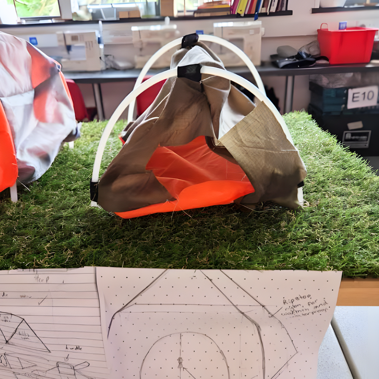 Student prototype of a portable shelter model using a curved frame and fabric covering, displayed on a grass base with annotated design sketches