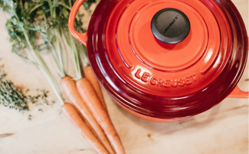 The image shows a red Le Creuset Dutch oven filled with carrots and garnished with fresh herbs.