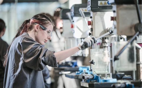 The image shows a person working on a machine in a factory setting, surrounded by industrial equipment.