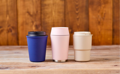 The image shows three reusable travel mugs in different colours arranged on a wooden table.