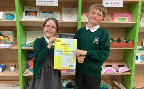 Two primary school children smiling and holding a D&T Mark certificate in front of a classroom display of student projects.