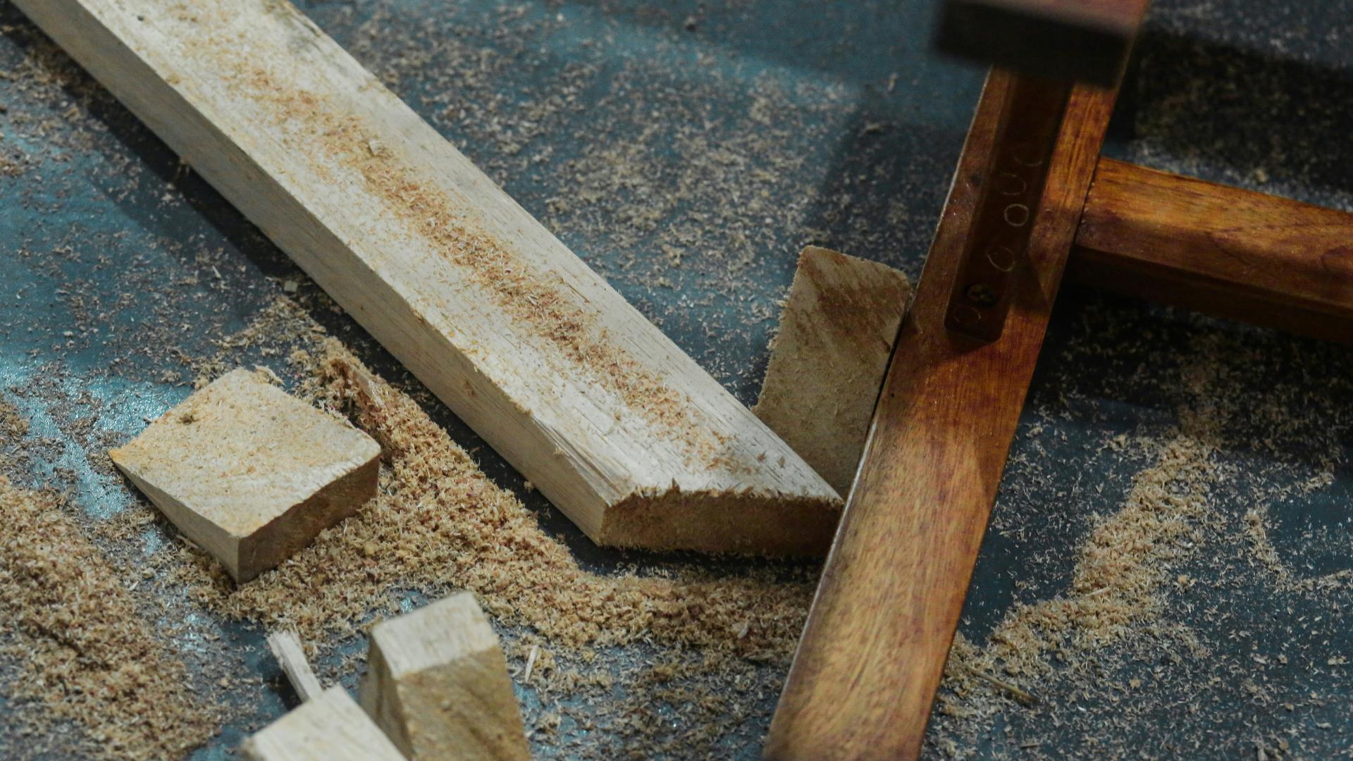 Close-up of a woodworking scene showing various pieces of cut wood, sawdust scattered on the ground, and part of a wooden structure or furniture frame.